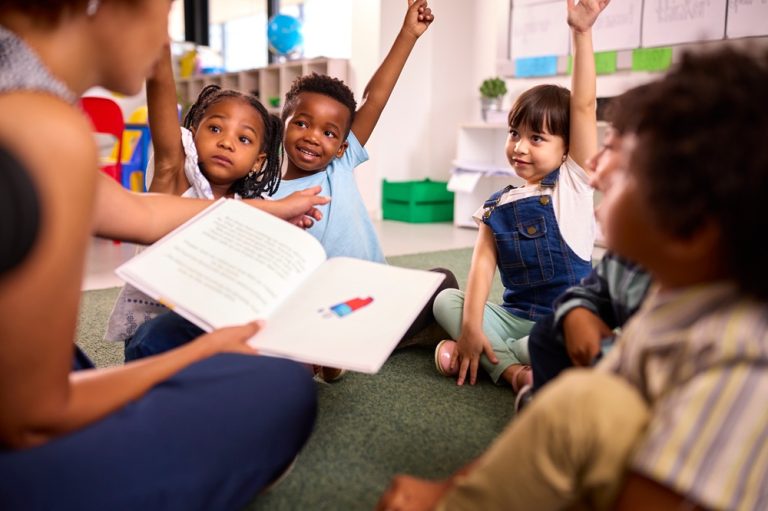 Female Teacher Reads To Multi-Cultural Elementary School Pupils Sitting On Floor In Class At School